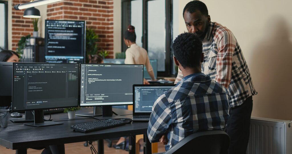 software developer writing code on laptop and computer keyboard looking at multiple screens with programming language is interrupted by coworker asking for advice