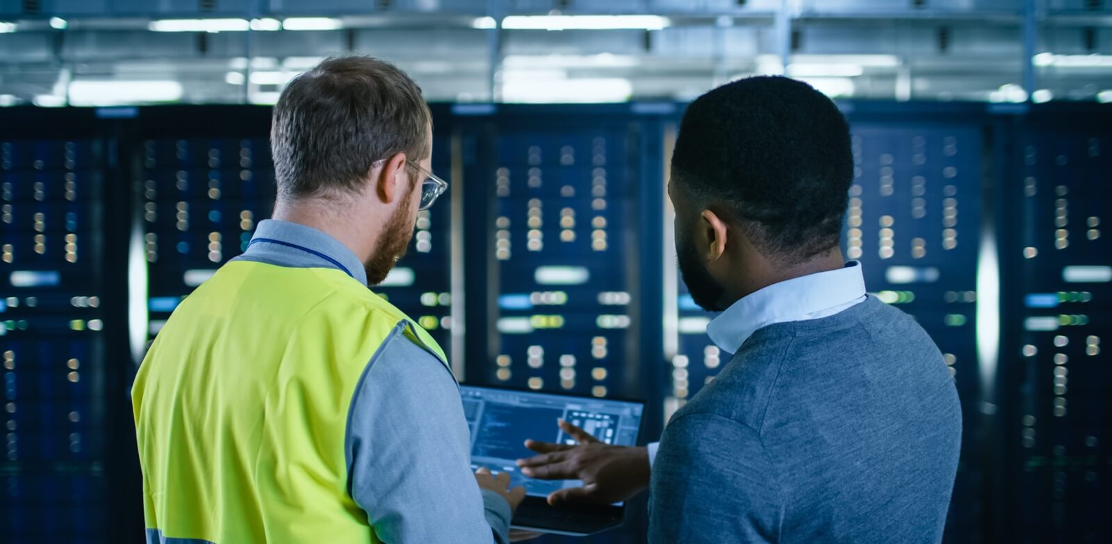 Back View on Bearded IT Engineer in Glasses and High Visibility Vest with a Laptop Computer and Black Technician Colleague Talking in Data Center while Standing Next to Server Racks (1)