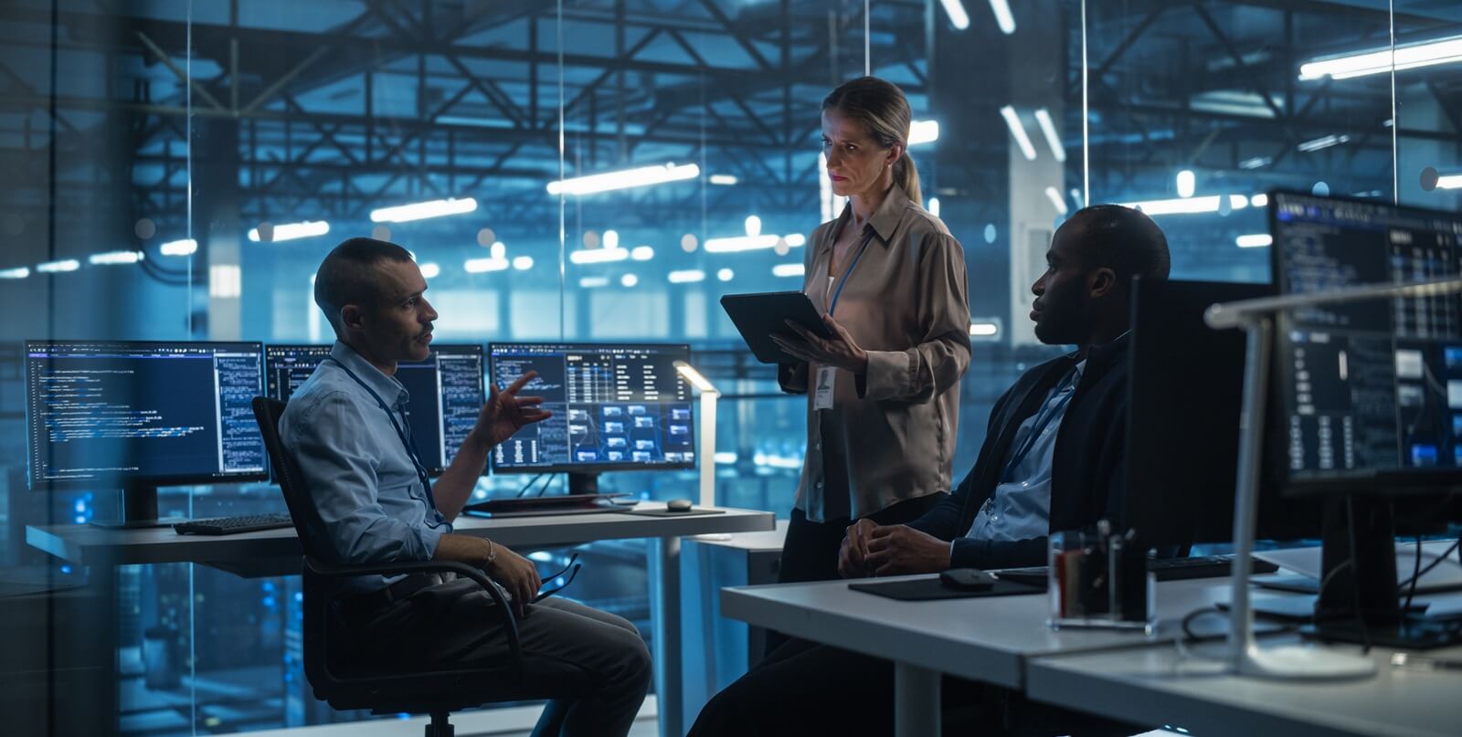two young male sitting down and female standing with a tablet computer exchange ideas in the data center office