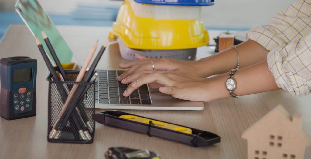 hand professional architect, engineer or interior hands working with laptop and safety helmet on workplace desk in office center at construction site